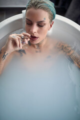A young sexy tattooed girl with a cigar enjoying moments of hedonism while having a bath in the bathroom. Relaxation, bath, bathroom, home