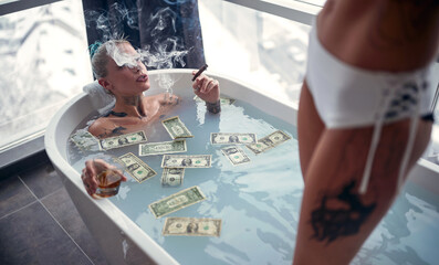 A young sexy tattooed lesbian couple enjoying a bath in the bathroom with cigars and a drink and...