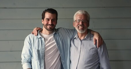 Happy young grown up son embracing shoulders of smiling proud middle aged 60s father, looking together at camera. Emotional positive two male generations family showing strong sincere relationships.