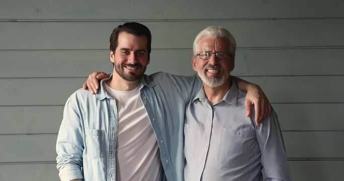 Happy young grown up son embracing shoulders of smiling proud middle aged 60s father, looking together at camera. Emotional positive two male generations family showing strong sincere relationships.