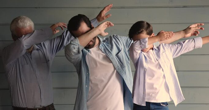 Playful young man doing dab gesture with joyful middle aged old father and small kid son, standing near wall. Funky happy multigenerational family presenting popular internet meme pose together.