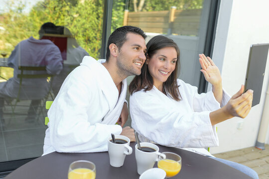 Happy young couple in bathrobes having breakfast outdoors while they are on doing a video call