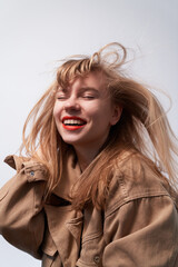 Obraz na płótnie Canvas Young fashion sensual woman laughing and posing on gray wall background dressed in hipster style beige jacket, with long hair and red lipstick