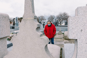 Man wearing a mask, sad and praying, in front of the grave of a deceased relative in times of covid-19