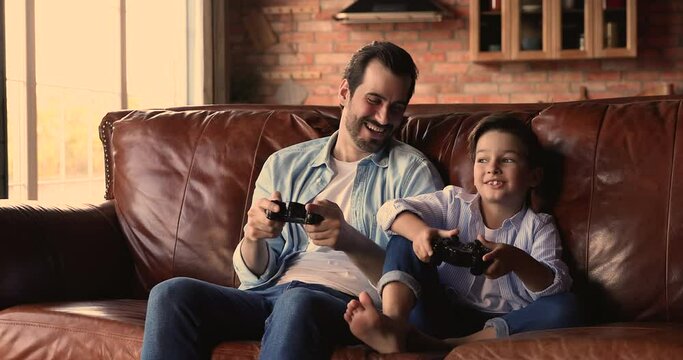Overjoyed emotional happy sincere young father using remote controllers, playing video games with laughing small school aged kid son in living room, weekend leisure pastime, domestic activity.