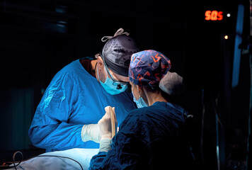 Surgeon performing cosmetic surgery in hospital operating room. Surgeon in mask wearing loupes...