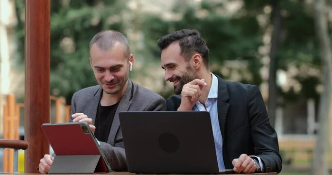 The male partners are discussing the project, looking at the tablet and smiling, applauding. Two men work on laptops in a cafe on the street.