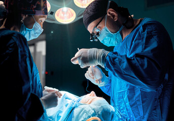 Surgeon performing cosmetic surgery in hospital operating room. Surgeon in mask wearing loupes...