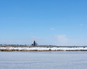 woman on bicycle in dutch winter landscape with frozen canal rides on dike