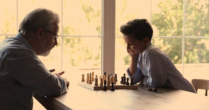Concentrated pleasant older mature grandfather teaching small grandson playing wooden board game, moving chess, enjoying leisure weekend playtime together at home, intellectual brainwork concept.