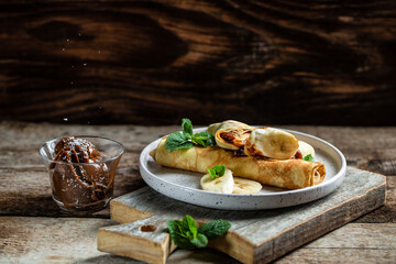 Crepes with chocolate and banana, rolled on white plate. Thin pancakes, blini. Sweet dessert. Top view with copy space