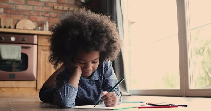 Full length interested small adorable african american curly girl lying on warm heated wooden floor, drawing alone in paper album, developing creative art skills in kitchen, domestic children activity