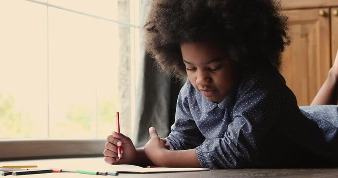Adorable small biracial african ethnicity happy kid girl lying on warm floor, entertaining alone drawing pictures in paper album, developing creativity art skills, enjoying free leisure hobby playtime