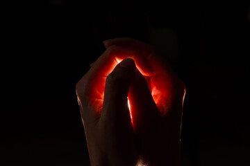 Two hands folded squeeze the light inside, reddish light inside the hands, black background. Origin of the idea, concept