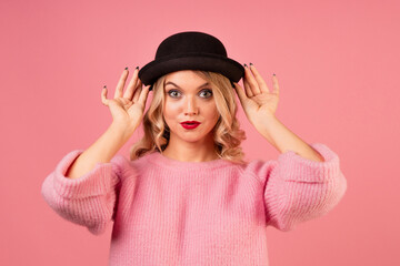A young, attractive woman in a warm pink sweater and a black bowler hat on a pink background, blonde with curly hair and red lipstick