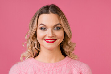 Fototapeta premium Young attractive woman in a warm pink sweater on a pink background, blonde with curly hair and red lipstick