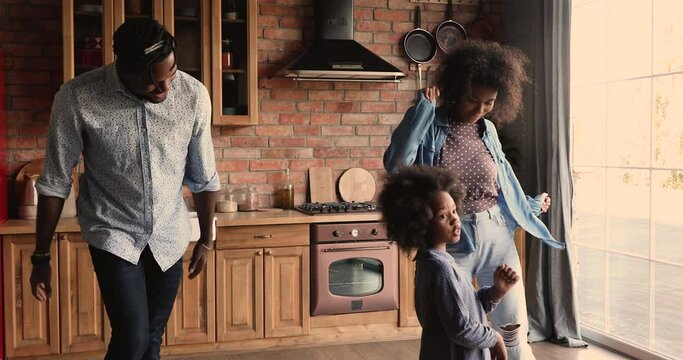 Small cute happy energetic african american kid girl dancing in kitchen with smiling couple parents, enjoying carefree weekend or celebrating moving into new big own house, family activity concept.