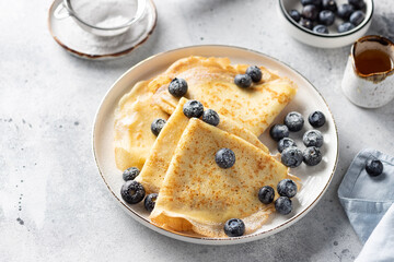 Homemade crepes pancakes served with blueberries and powdered sugar on white plate. Tasty breakfast food. Gray background. side view