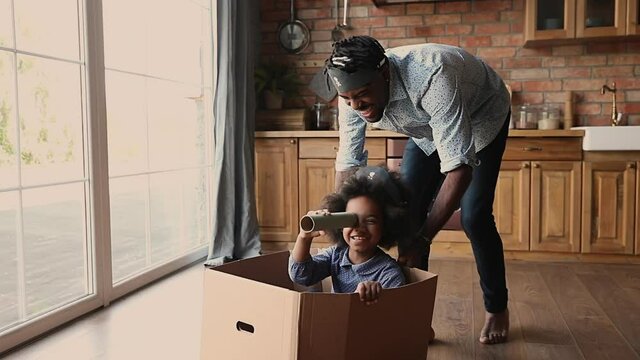 Joyful young african american father playing pirates with adorable small kid daughter sitting in box, imagining ship captain. Laughing little biracial child girl having fun with caring daddy indoors.