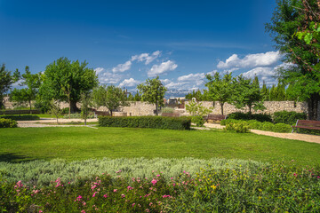 Small city park with benches, flowers, green hedges, trees and grass. Nin town cemetery and mountain range of Dinaric Alps in background, Croatia
