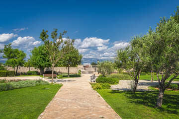 Small city park with stone footpath, benches and green trees and grass in Nin town, cemetery and mountain range of Dinaric Alps in background, Croatia