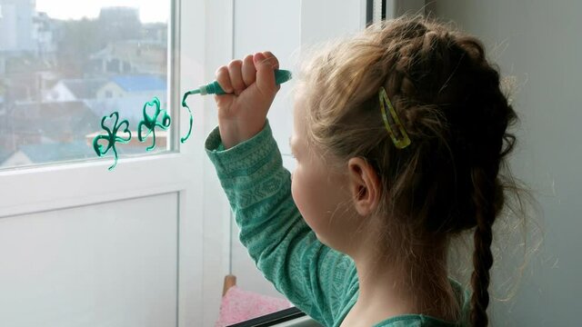 Drawing St. Patrick's Day Child painting green three-leaved shamrocks indoor, festive home decoration, quarantine family leisure. Little girl draws clover on window glass. Stay home concept New normal