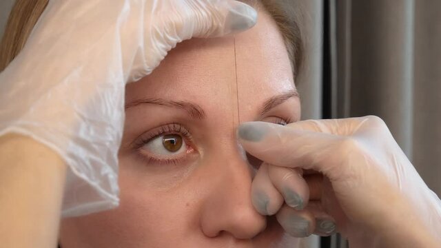 Correction and shaping procedures of female eyebrows in beauty salon. Professional styling of client face and stylist's hands working with special thread to mark new contour of brows close up