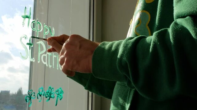 Drawing St. Patrick's Day Man painting green three-leaved shamrocks indoor, festive home decoration, quarantine family leisure. Father draws clover leaves on window glass. Stay home concept New normal