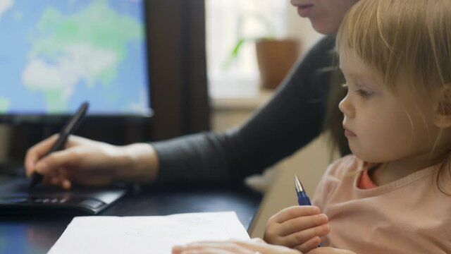 Online working during lockdown with a child, mommy and baby are drawing. Tender woman hand uses tab and a digial pen, kids hands writing on a paper. Cute interested blond girl by the computer table