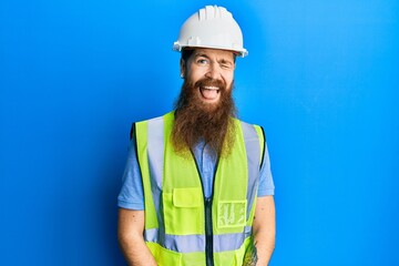 Redhead man with long beard wearing safety helmet and reflective jacket winking looking at the camera with sexy expression, cheerful and happy face.