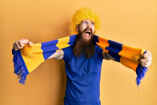 Redhead man with long beard football hooligan cheering game wearing funny wig angry and mad screaming frustrated and furious, shouting with anger. rage and aggressive concept.