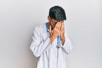 Young handsome man wearing doctor uniform and stethoscope with sad expression covering face with hands while crying. depression concept.