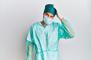Fototapeta na wymiar Young handsome man wearing surgeon uniform and medical mask confuse and wonder about question. uncertain with doubt, thinking with hand on head. pensive concept.