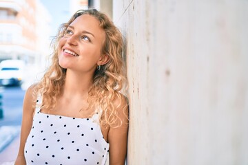 Naklejka premium Young beautiful caucasian woman with blond hair smiling happy outdoors on a summer day leaning on the wall