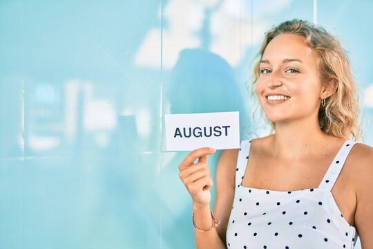 Young beautiful caucasian woman with blond hair smiling happy outdoors on a summer day holding August word