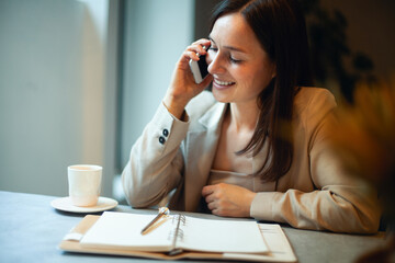 Young woman with long dark straight hair in in a beige nude business suit is talking on phone and writing notes