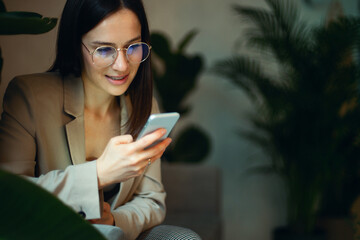 Beautiful businesswoman in suit sitting on sofa and reading text message on mobile phone with smile