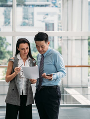 Two business people reading papers together in modern office with big windows.
Two serious business people standing and talking about documents in office lobby