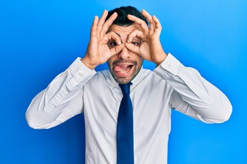 Young hispanic man wearing business clothes doing ok gesture like binoculars sticking tongue out, eyes looking through fingers. crazy expression.