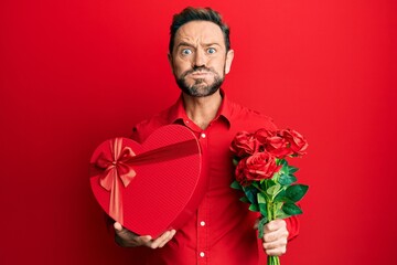 Middle age man holding anniversary present and bouquet of flowers puffing cheeks with funny face....