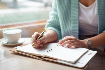 Well groomed woman hand holding gold pen and writing notes with gold pen in notebook beside window.
