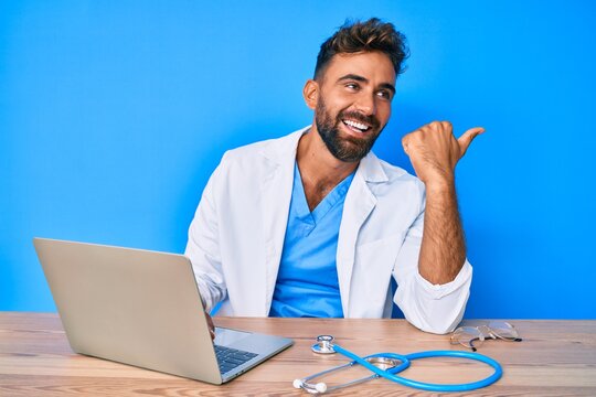 Young hispanic man wearing doctor uniform working at the clinic pointing thumb up to the side smiling happy with open mouth