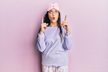 Beautiful middle eastern woman wearing sleep mask and pajama amazed and surprised looking up and pointing with fingers and raised arms.