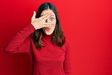 Young brunette woman wearing turtleneck sweater peeking in shock covering face and eyes with hand, looking through fingers afraid