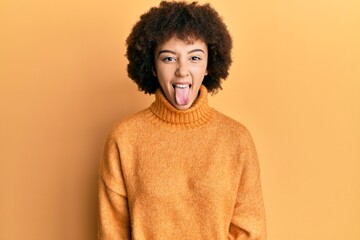Fototapeta na wymiar Young hispanic girl wearing wool winter sweater sticking tongue out happy with funny expression. emotion concept.