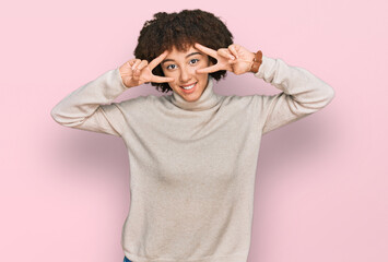 Young hispanic girl wearing wool winter sweater doing peace symbol with fingers over face, smiling cheerful showing victory