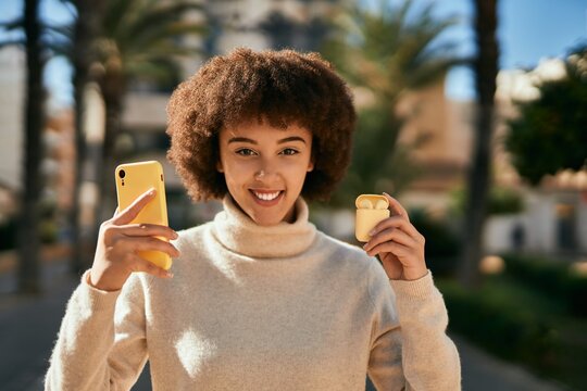 Young hispanic girl smiling happy using smartphone and holding earphones at the city.