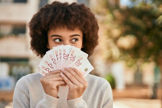 Young hispanic girl smiling happy holding israel shekels covering mouth at the city.
