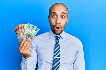 Hispanic adult man holding australian dollars scared and amazed with open mouth for surprise, disbelief face