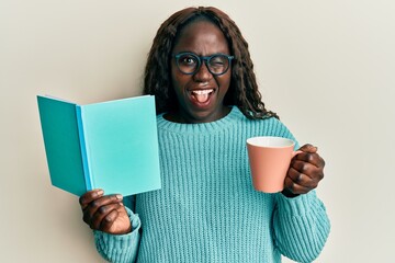 African young woman reading a book and drinking a cup of coffee winking looking at the camera with...
